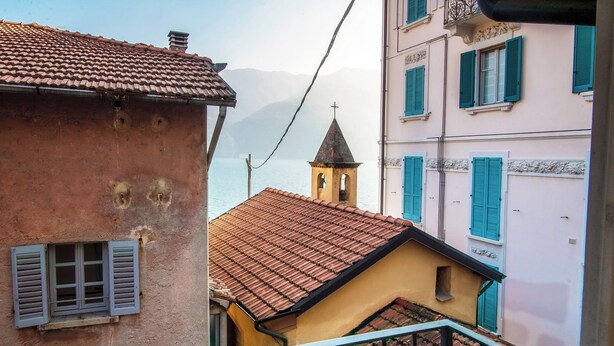 A narrow street with buildings featuring red-tiled roofs and blue shutters. A small bell tower is visible in the distance. - Lake Nest (Carate Urio)