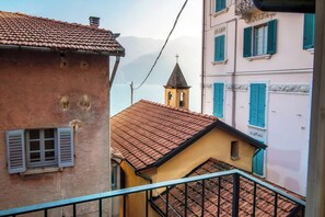 Rua estreita com edifícios com telhados vermelhos e persianas azuis. Uma pequena torre de sino é visível ao longe.