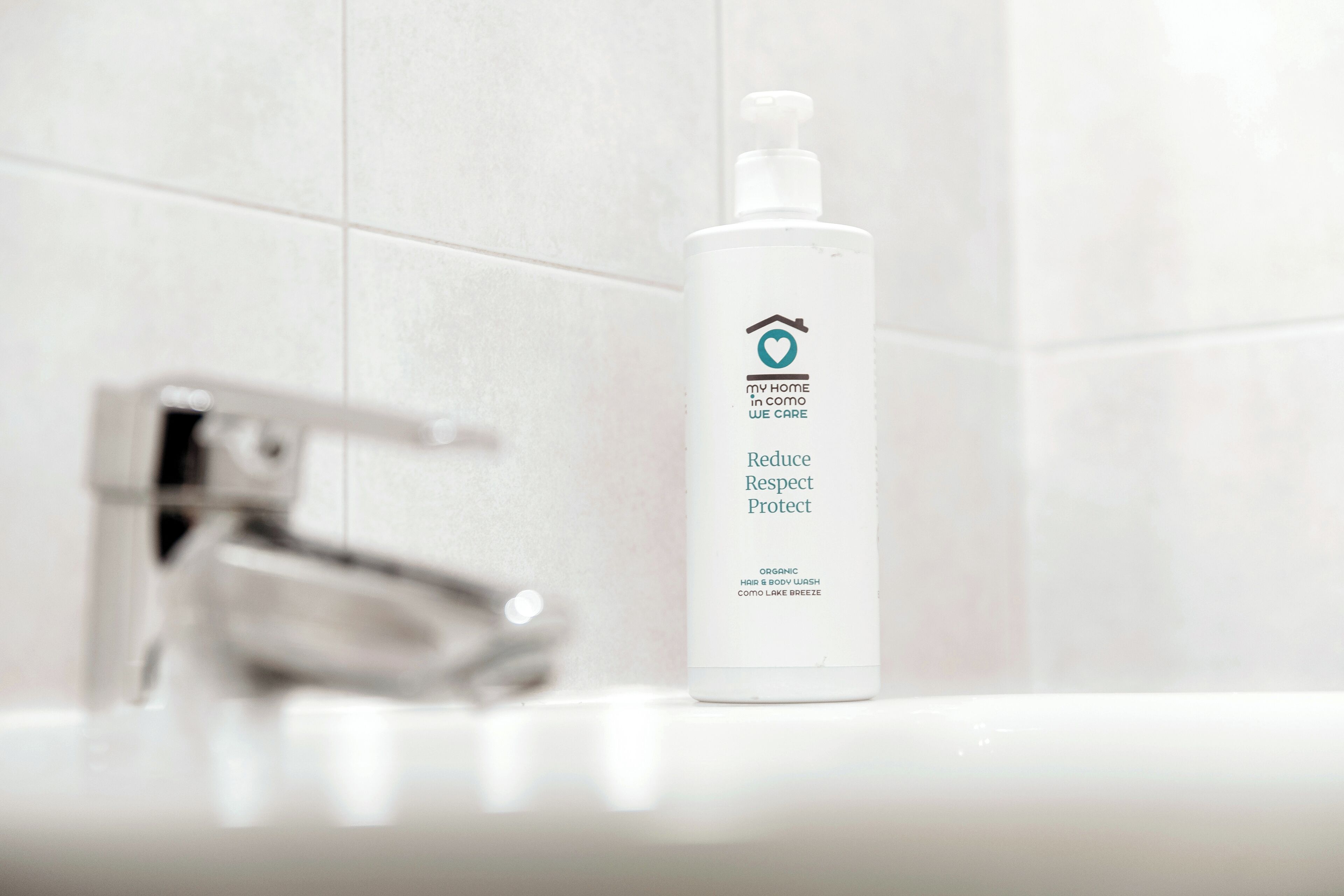 Une salle de bain avec un lavabo blanc, un robinet chromé et une bouteille de gel douche biologique pour les cheveux et le corps.