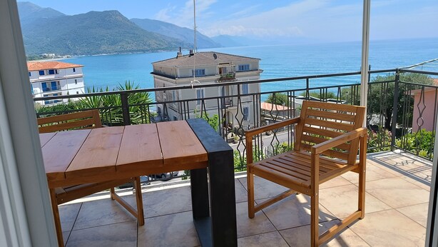 Ein Balkon mit Holztisch und Stuhl, der Blick auf eine Küstenlandschaft mit Gebäuden und Bergen bietet.