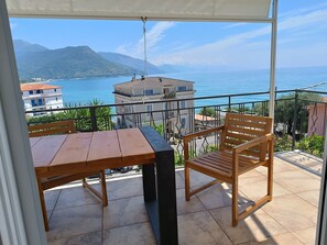 Ein Balkon mit Holztisch und Stuhl, der Blick auf eine Küstenlandschaft mit Gebäuden und Bergen bietet.