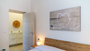 Ein Schlafzimmer mit Holz-Kopfteil, einem Bett mit floralem Bettzeug, einem an der Wand hängenden abstrakten Gemälde, einer Nachttischlampe und Blick in ein Badezimmer mit rundem Spiegel und Ablagefläche mit kleinen Pflanzen.