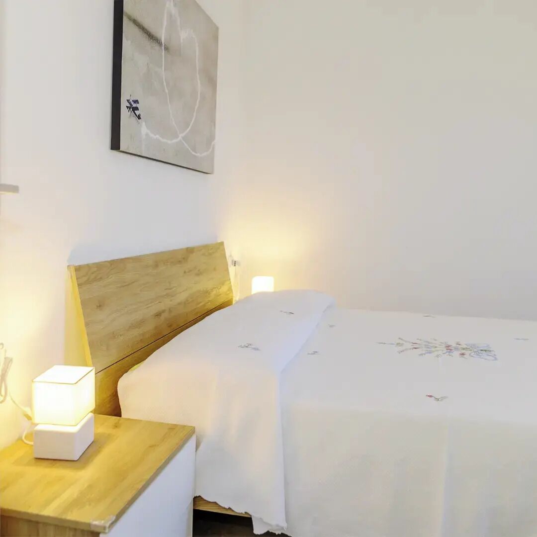 Een net opgemaakt bed met witte lakens, een houten hoofdbord en een nachtkastje met een lamp.