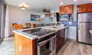 A modern kitchen with a central island, stainless steel appliances, wooden cabinets, and a dining area with a view. - Kamaole Nalu, #504^ 2 Bedroom Condo by RedAwning (Kihei)