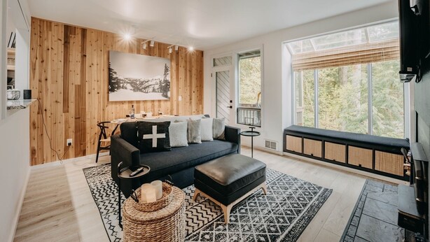 Ein modernes Wohnzimmer mit einer Wandpaneelaus Holz, einem schwarzen Sofa mit Kreuzkissen, einem schwarzen Hocker, einem gemusterten Teppich und einem großen Fenster mit Blick auf Bäume.
