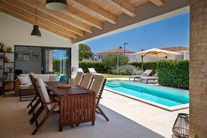 A modern outdoor living area with a pool, wooden furniture, and a glass wall overlooking a garden.