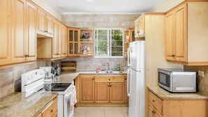 A kitchen with wooden cabinets, a white refrigerator, a microwave, and a window with a view of greenery. - NEW Gated Beach front Condo (Nassau)