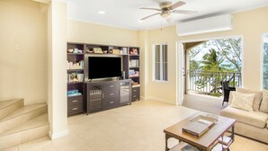 A spacious living room with a flat-screen TV, a bookshelf, a coffee table, and a balcony with a view of palm trees. - NEW Gated Beach front Condo (Nassau)