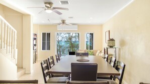 A spacious dining area with a long table, multiple chairs, ceiling fans, and large windows offering a view of trees. - NEW Gated Beach front Condo (Nassau)
