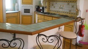 A kitchen with yellow cabinets, a tiled backsplash, a stove, and a bar area with stools. - Maison Avec Piscine Dans le Parc du Luberon, Chemin de Randonnée à Proximité (Gargas)