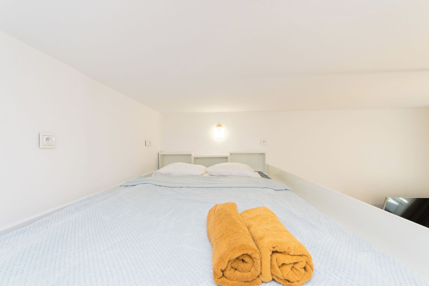 A single bed with a light blue bedspread, two white pillows, and two rolled-up towels on the bed.