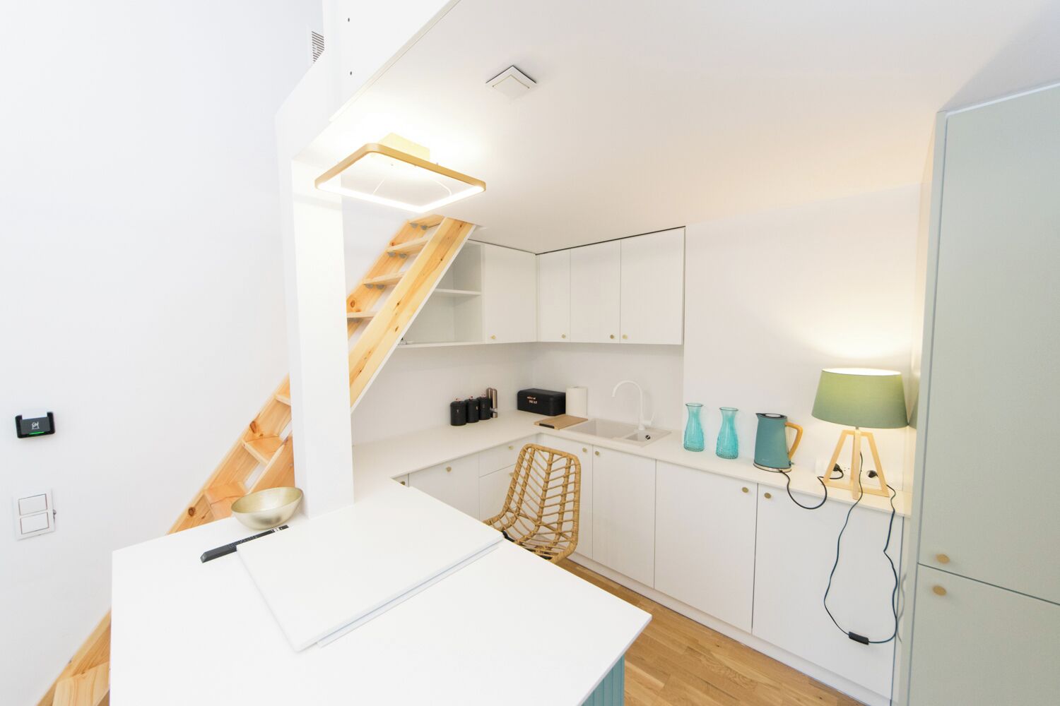 Une cuisine compacte avec des meubles blancs, un escalier en bois, un plan de travail blanc et une petite table à manger.