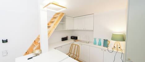 Une cuisine compacte avec des meubles blancs, un escalier en bois, un plan de travail blanc et une petite table à manger.