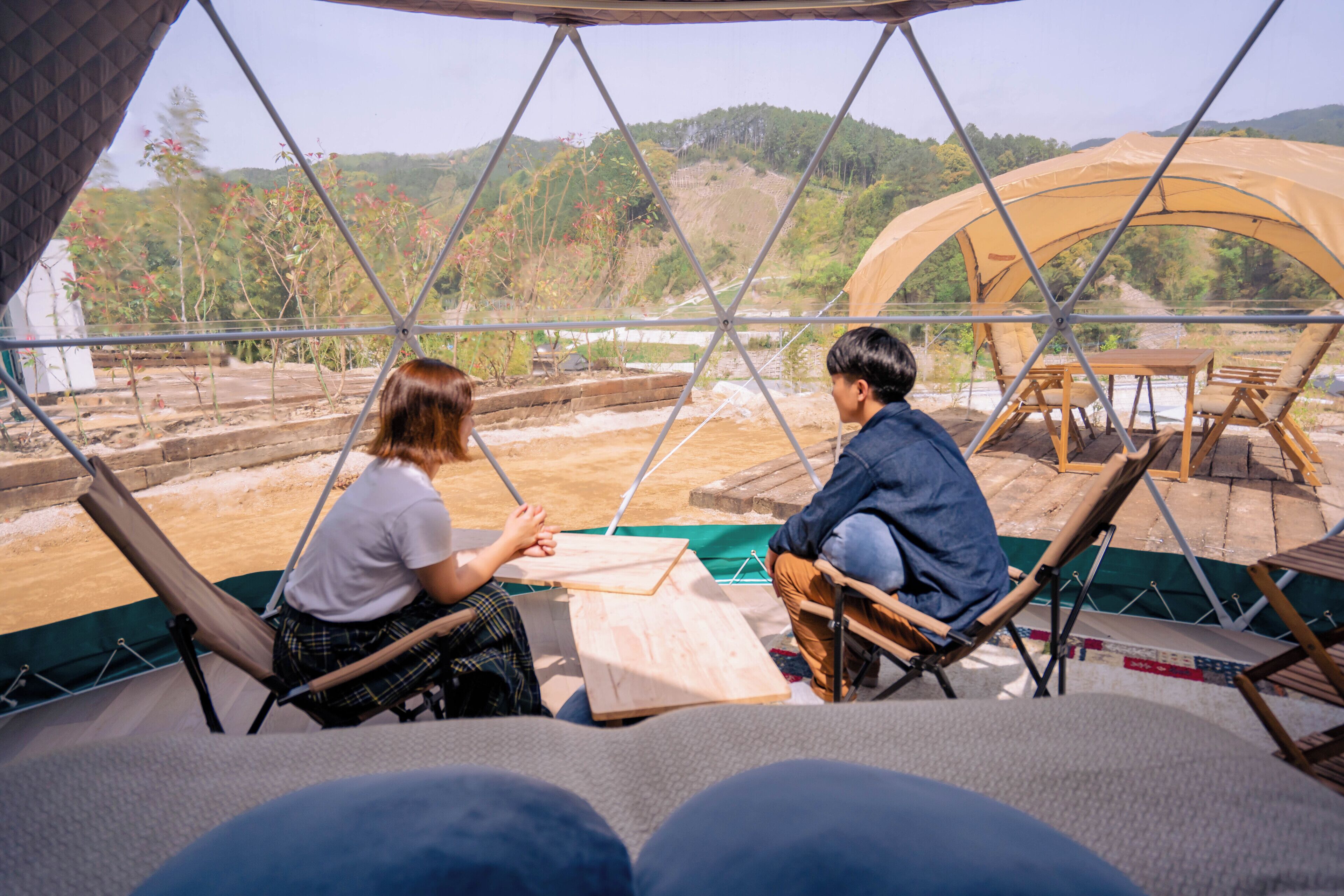 Two people seated at a table inside a geodesic dome structure with a view of a hilly landscape.