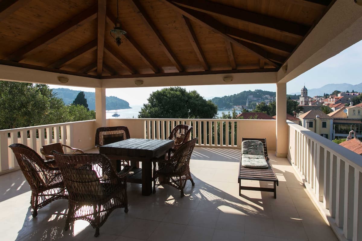 A covered outdoor patio with a table and chairs, overlooking a scenic view of a body of water and hills.