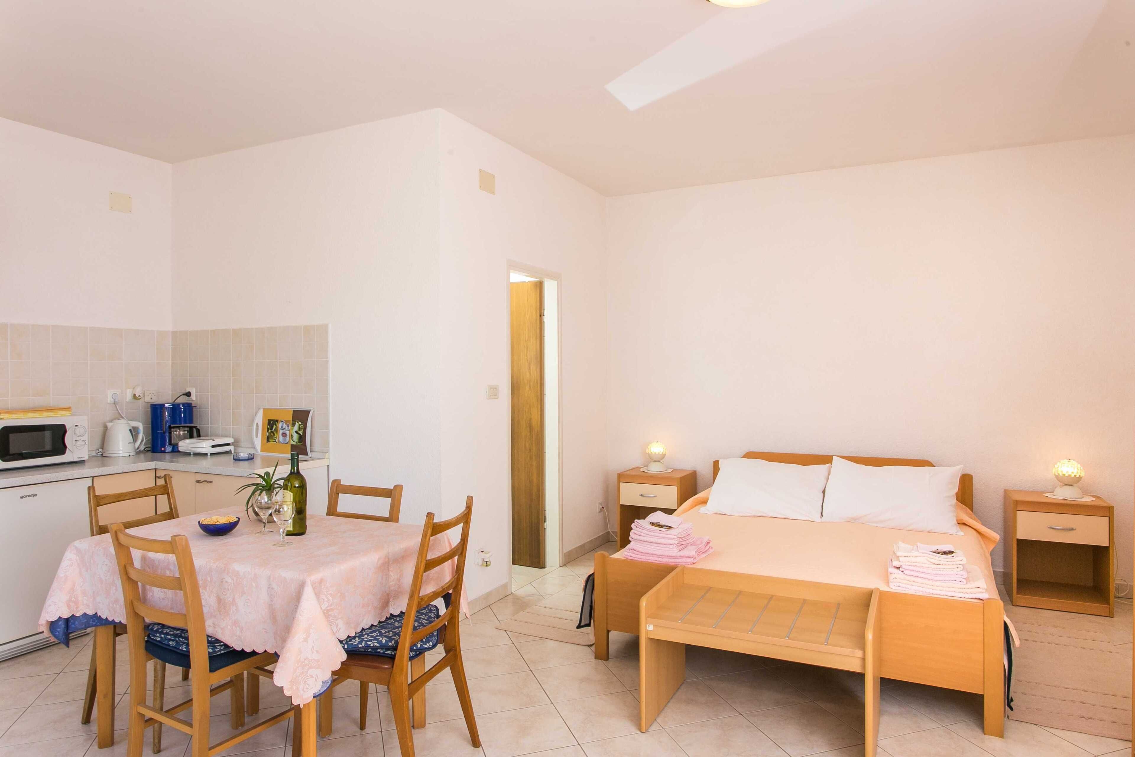 Une chambre avec un lit en bois, des tables de chevet et une petite lampe de chevet. On y trouve également un coin repas avec une table et des chaises, ainsi qu’une kitchenette équipée d’un four à micro-ondes et d’une bouilloire.