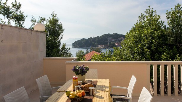 A table set with fruits, a flower vase, and a croissant on a balcony overlooking a coastal town. - Apartments Miljan Popovic (Konavle)
