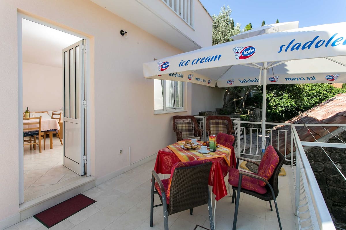 An outdoor patio with a table set for two, covered by a large ice cream umbrella, and a door leading to an interior dining area.