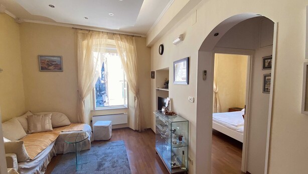 A hotel room with a sofa, a small round table, a glass cabinet with items, a window with curtains, and a bed.
