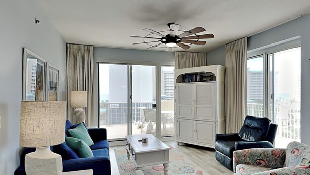 A modern living room with a blue sofa, a white coffee table, a black armchair, and a patterned ottoman. There is a ceiling fan, a large window with curtains, and a view of the city. - Ariel Dunes #510 | Gulf Side Condo, Amazing Water Views + Resort Amenities! (Miramar Beach)