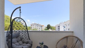 A balcony with two wicker chairs, one hanging and one seated, a small round table with a potted plant, and a view of residential buildings in the background. - Brand new apartment Excellence with 3 bedrooms and 3 bathrooms (Pula)