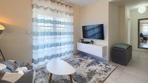 A modern living room with a flat-screen TV, a white sofa, a patterned rug, and a large window with sheer curtains. - Brand new apartment Excellence with 3 bedrooms and 3 bathrooms (Pula)