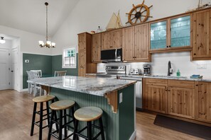 A kitchen with wooden cabinets, a central island with stools, a microwave, and a ship wheel decoration. - Captain and Crew at Seaside Beach (Seaside)