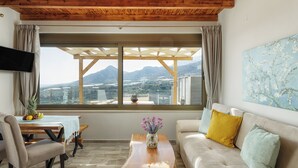 A living room with a wooden ceiling, a sofa with cushions, a coffee table with a vase, a TV on the wall, and a window with a view of mountains. - 2 Bedroom Villa in Mediterranean View Villas in Makri gialos (Makry Gialos)