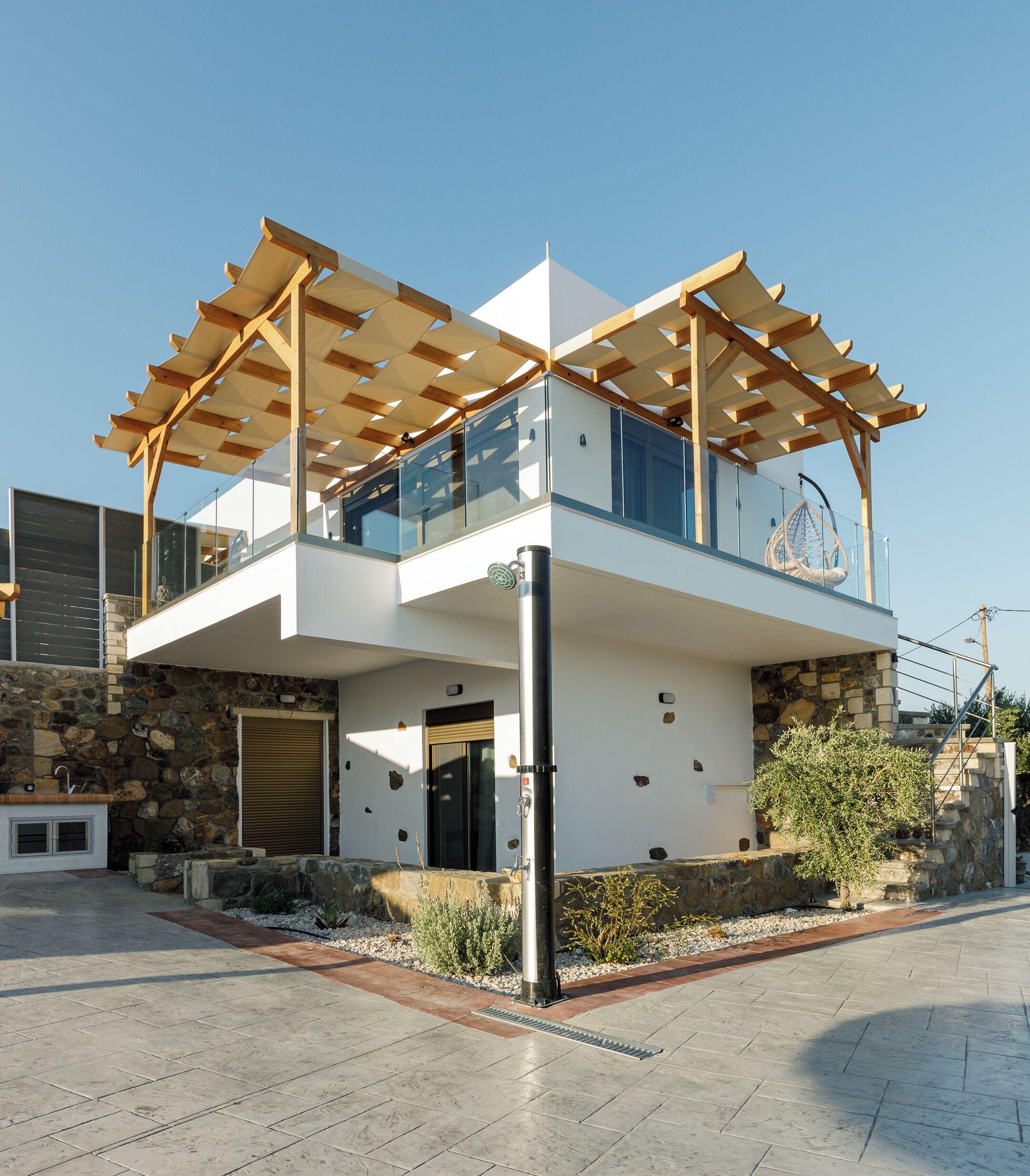 Une maison moderne à deux étages, dotée d’une façade en pierre, d’un balcon agrémenté d’une pergola en bois et d’un fauteuil suspendu.