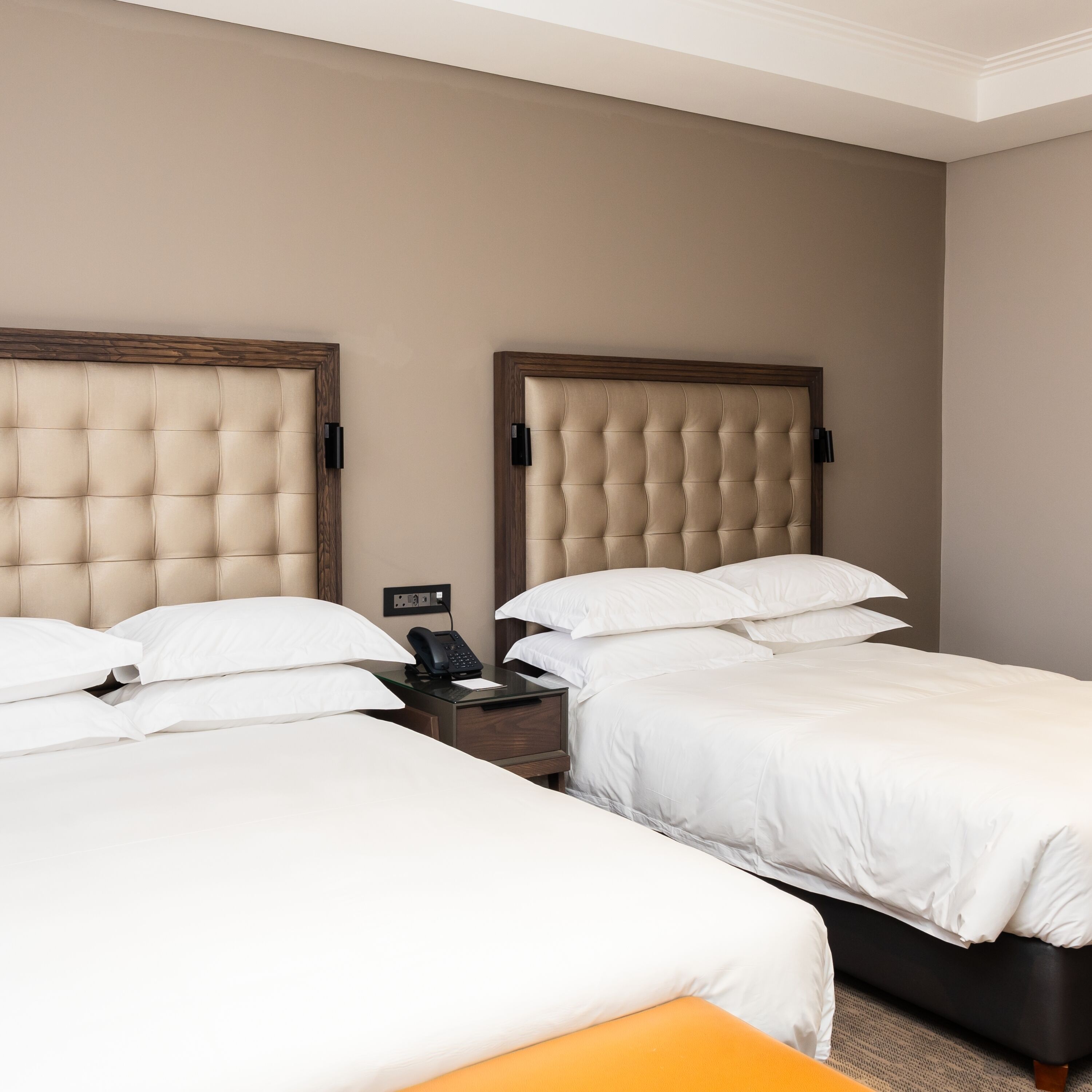 A hotel room with two beds, each with a wooden headboard and white bedding. There is a bedside table with a phone and a small clock.
