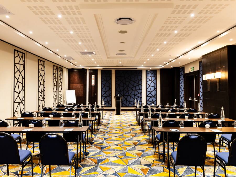 Een congreszaal met een vloer met een geometrisch patroon, rijen tafels met stoelen en een modern plafondontwerp.