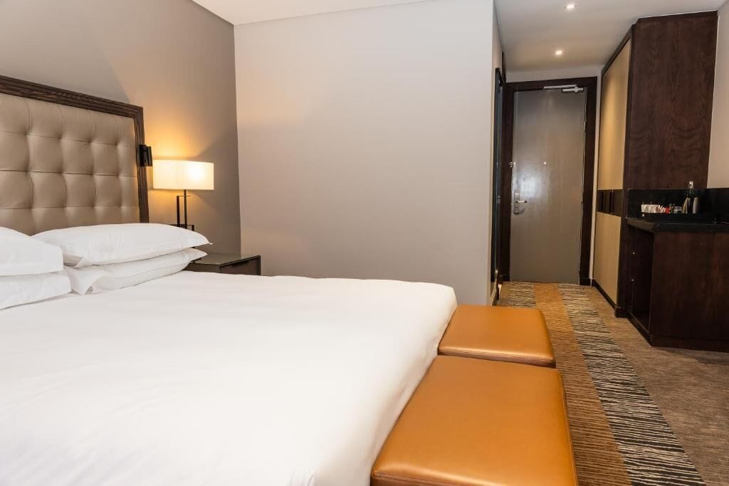 A hotel room with a large bed, a nightstand with a lamp, a wooden wardrobe, and a bench.