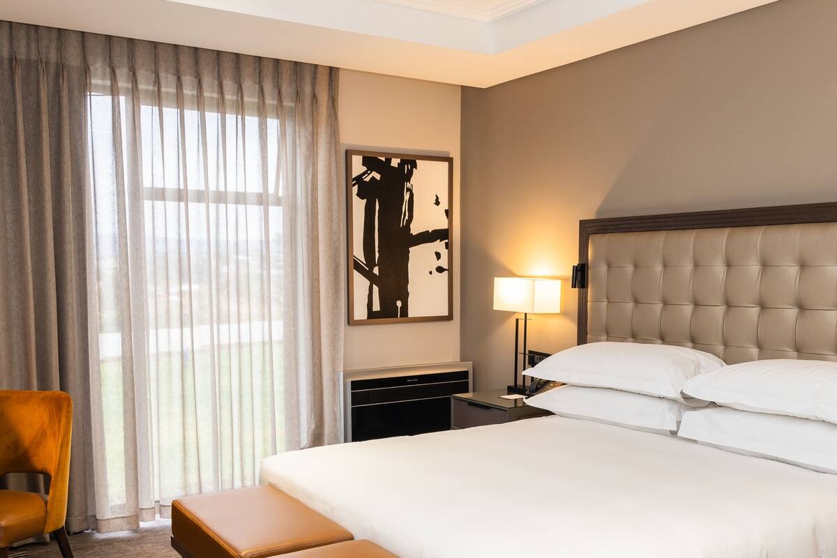 A hotel room with a large bed, a bedside table with a lamp, a chair, and a large window with curtains.