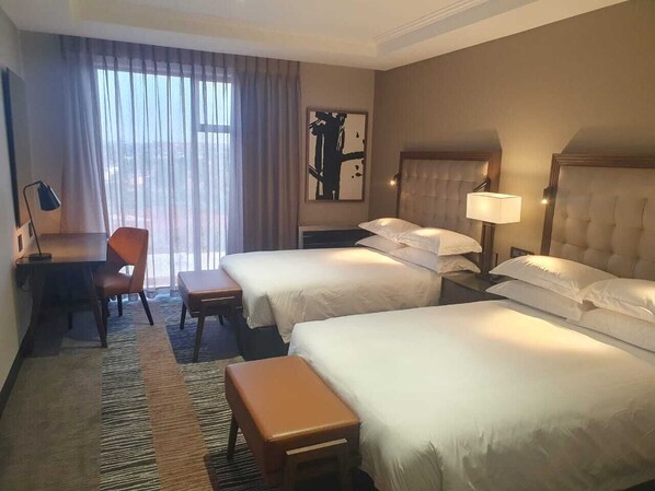 Habitación de hotel con dos camas, un escritorio, una silla y un ventanal con cortinas.