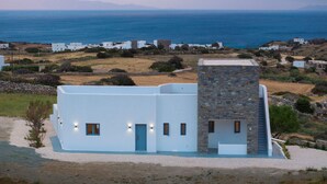 Un moderno edificio blanco con una torre de piedra, rodeado por un paisaje costero con casas al fondo.