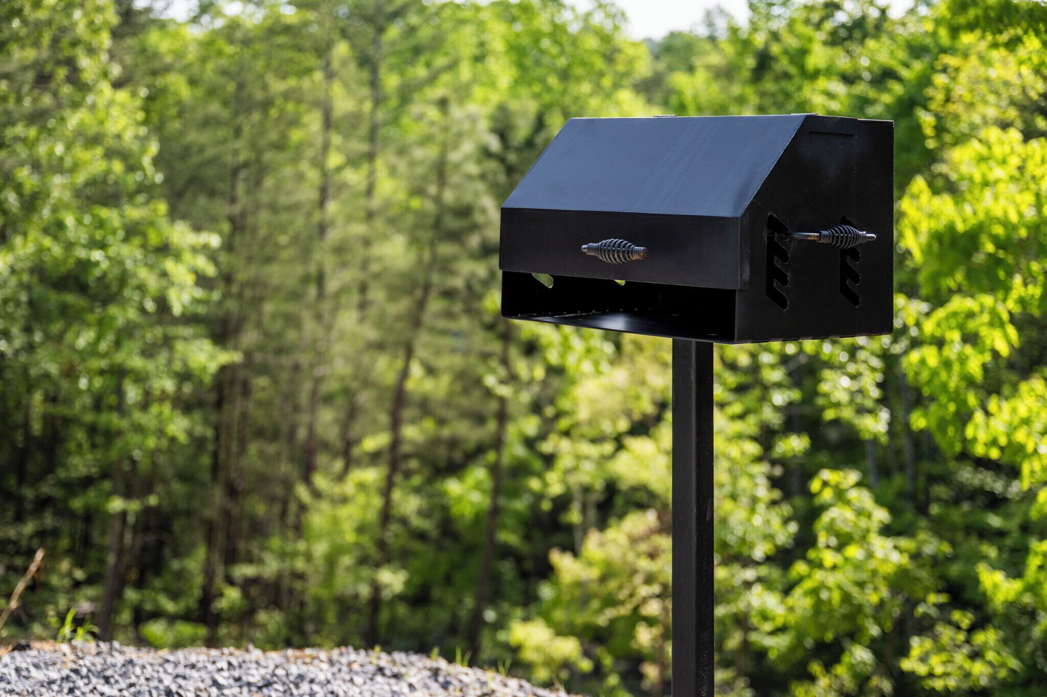 A black barbecue grill with a ribbed handle and a vent, set against a backdrop of dense green foliage.