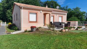 Une petite maison avec un toit en tuiles, une terrasse avec des meubles d’extérieur et un jardin fleuri.
