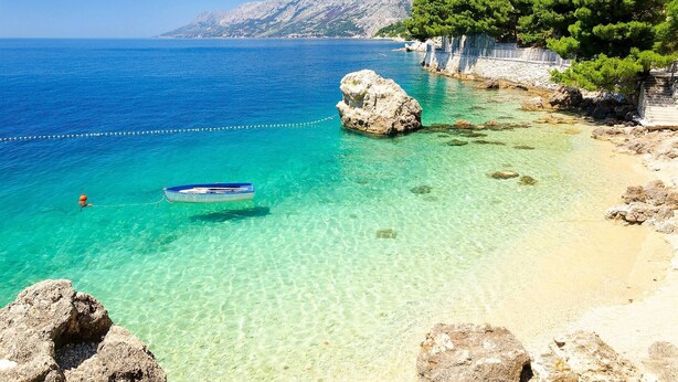 A coastal scene with clear turquoise waters, a small boat anchored near a rocky outcrop, and a building with balconies overlooking the sea. - Adria View, Apartment Dado, with pool (Makarska)