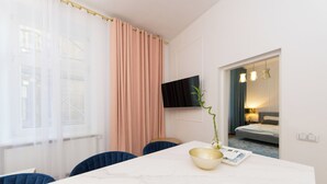 Une chambre d’hôtel moderne avec une table à manger en marbre, une grande fenêtre ornée de rideaux l égers, un téléviseur à écran plat fixé au mur et un lit doté d’une tête de lit et de lampes de chevet.