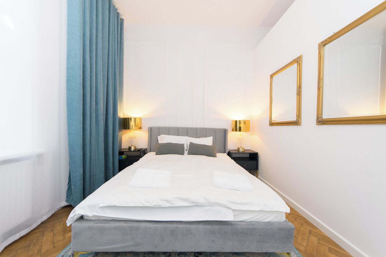 A neatly arranged hotel room with a bed, two bedside tables with lamps, a large mirror, and a window with blue curtains.