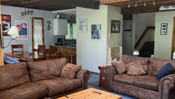 A spacious living room with two brown leather sofas, a wooden coffee table, and a patterned rug. A wooden railing overlooks the space, and a kitchen is visible in the background. - Roomy ski condo next to Mt. Snow, on the mover bus, with hot tub and sauna! (Dover)