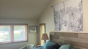A bedroom with a large bed, a headboard, a window with blinds, a bedside table, a lamp, and a wall tapestry. - Roomy ski condo next to Mt. Snow, on the mover bus, with hot tub and sauna! (Dover)