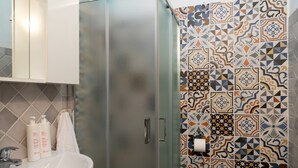 Une salle de bain avec un lavabo blanc, deux bouteilles de shampoing, une serviette et une douche dont les carreaux sont décorés de motifs.