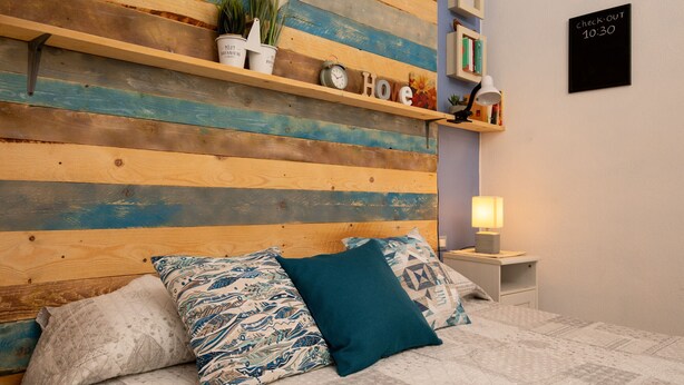 Une chambre à coucher avec une tête de lit en bois, un lit recouvert d’une literie à motifs, une table de chevet avec une lampe et une étagère ornée d’objets décoratifs.