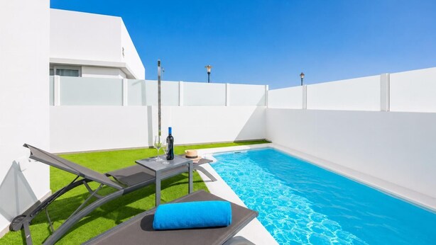 Un espace piscine moderne avec une piscine à l’eau d’un bleu limpide, une chaise longue, une table avec une bouteille et des verres, et un petit chapeau.
