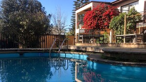A clear swimming pool with a wooden deck and a metal ladder, surrounded by trees and a building with red foliage.