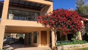 A two-story building with a covered patio area, a balcony with metal railings, and a large flowering bush in front.