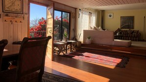 A spacious living room with a wooden ceiling, a sofa set, a dining table with chairs, and a view of red flowers through the glass door.
