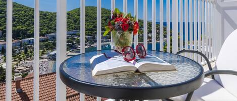 Ein runder Glastisch mit einem offenen Buch, einem Paar roter Brillen und einem Strauß roter Blumen auf einem Balkon mit Blick auf eine Küstenstadt und grüne Hügel.