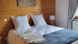 A bedroom with a wooden headboard, a bed with white bedding, a decorative sign, a small table with a lamp, and a window with curtains. - L'évasion au Chalet de Fraize Avec vue Imprenable,équipé de Vélos Électriques (Fraize)